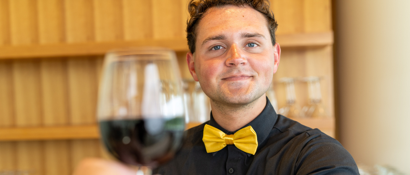 Person wearing black shirt and yellow bow tie and handing glass of wine